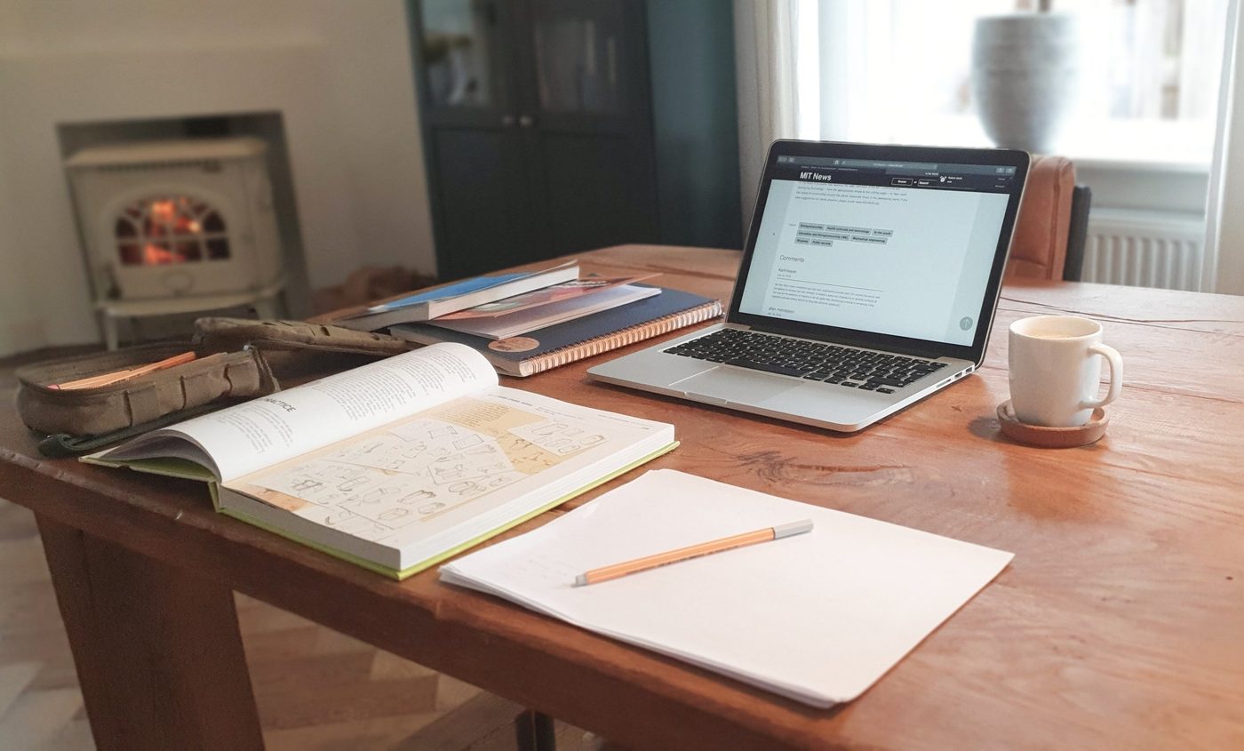 A laptop, coffee mug, various books and some paper with a pen sit atop a table in a home.